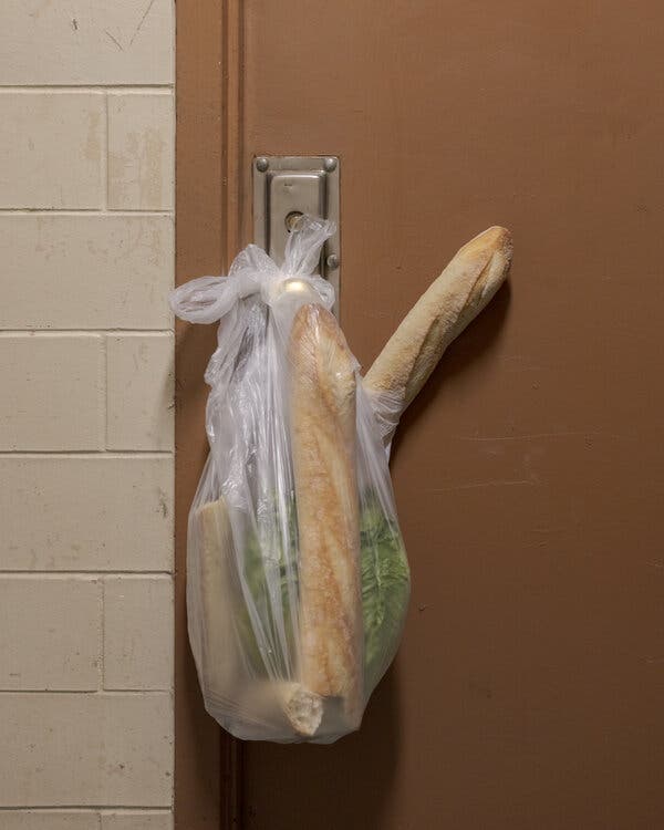 A plastic bag hanging from a doorknob holding French bread and vegetables.