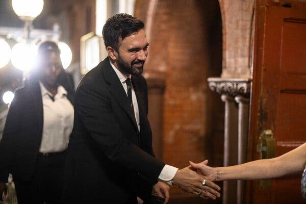 Zohran Mamdani, wearing a dark suit, smiles and reaches out to shake a person’s hand while entering a church.