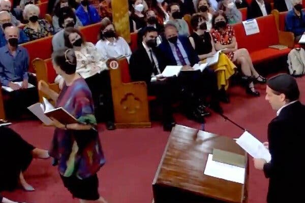 In a video screenshot, Zohran Mamdani and Brad Lander, both wearing suits and masks, sit in a pew among congregants inside a synagogue.