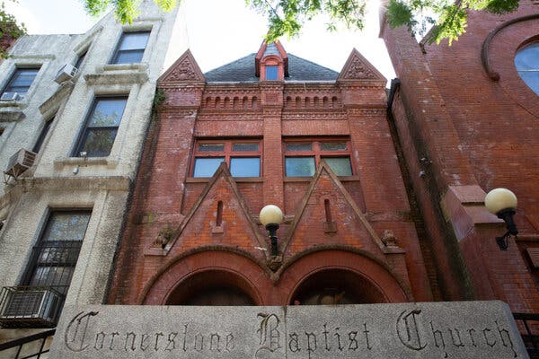 A brick building with arches on a city street, with a concrete sign in front of it reading “Cornerstone Baptist Church” in a Gothic font.